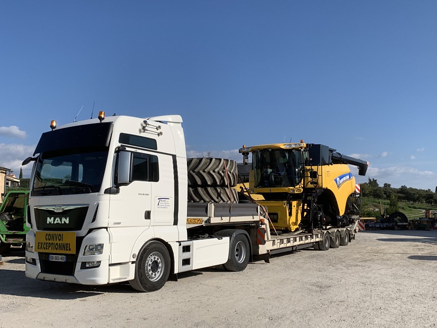 Un camion blanc MAN avec un panneau « Convoi exceptionnel » transporte une moissonneuse-batteuse jaune et de gros pneus sur une remorque, sur un terrain gravillonné.