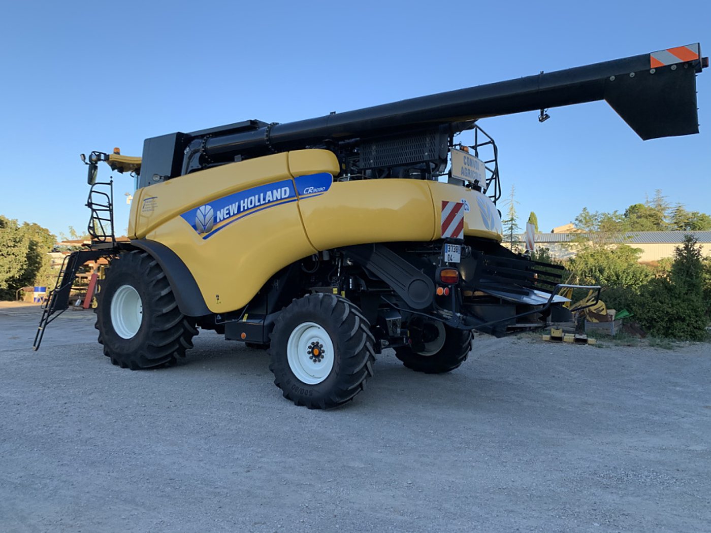 Une moissonneuse-batteuse New Holland jaune et noire, vue de l'arrière, est garée sur une cour en gravier sous un ciel bleu.